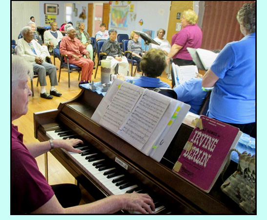 Photo was taken from behind Suzann while she is playing the piano, we can see the backs of Mary Kate and a few of the singers, and 9 of the audience members.
